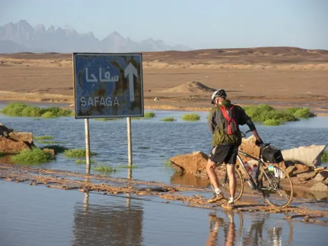 In this image we can see sign board, person, bicycle, water, stones, grass, sand, hills and sky.